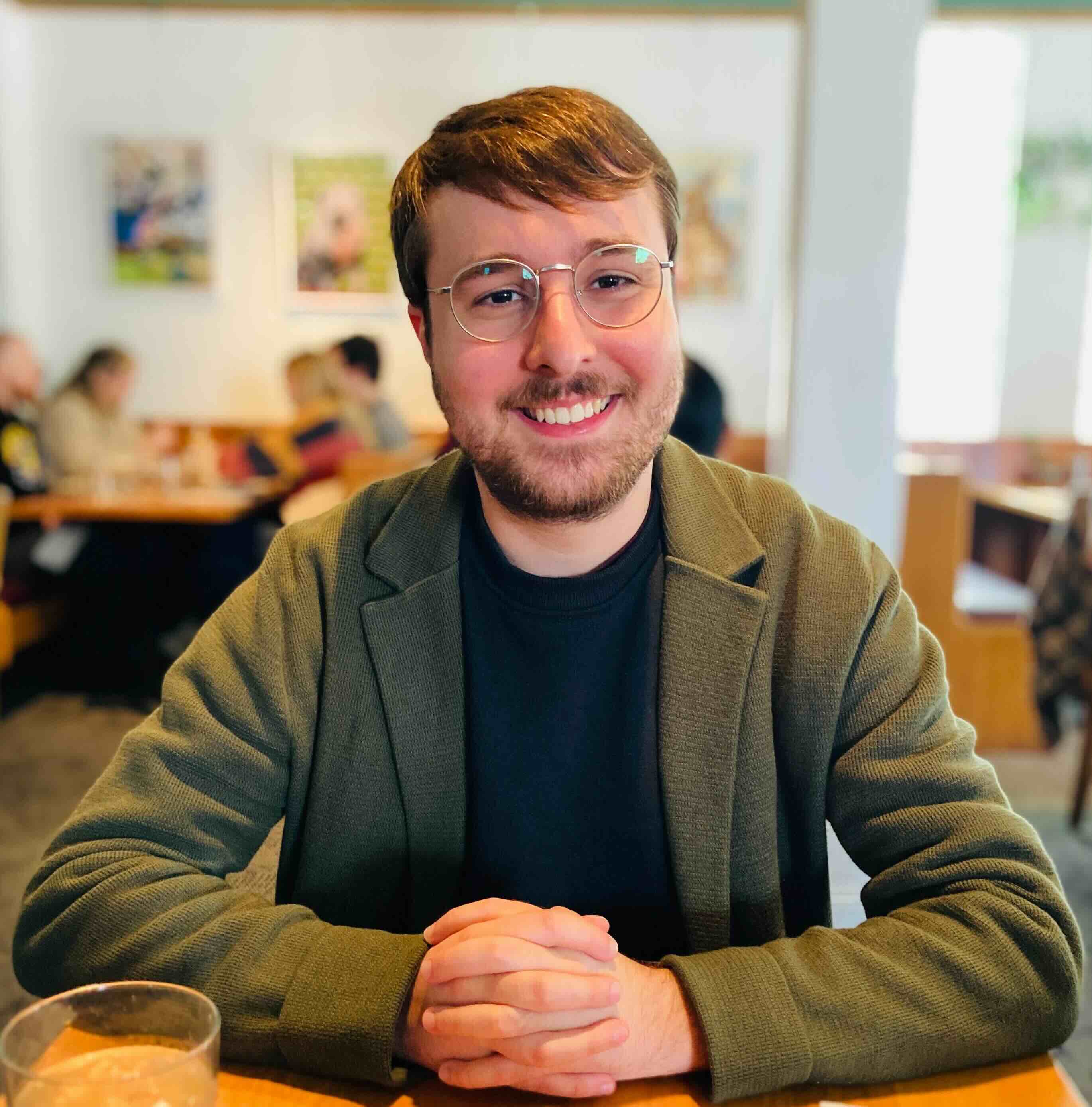 A picture of Mac Chaffee (pronounced CHAY-fee) sitting at a table while smiling with his hands clasped together in front of him.